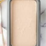 A rectangular metal baking pan filled with a smooth, light beige batter sits on a marble surface next to a striped towel and white bowls.