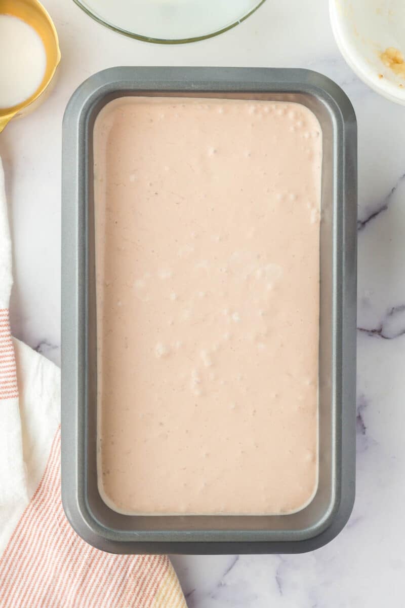 A rectangular metal baking pan filled with a smooth, light beige batter sits on a marble surface next to a striped towel and white bowls.