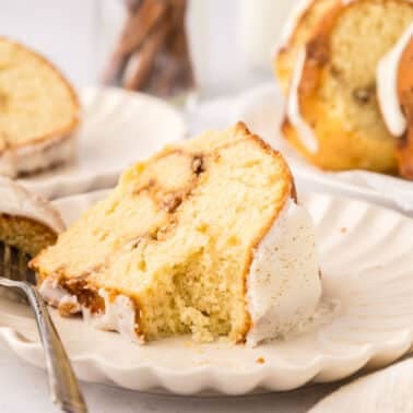 A slice of yellow bundt cake with white icing on a scalloped plate, partially eaten, with a fork resting on the plate and more cake in the background.