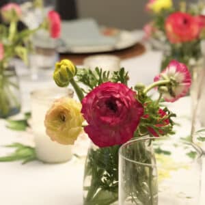 A small glass vase with pink, yellow, and white ranunculus flowers sits on a white tablecloth, surrounded by other floral arrangements and a white candle.