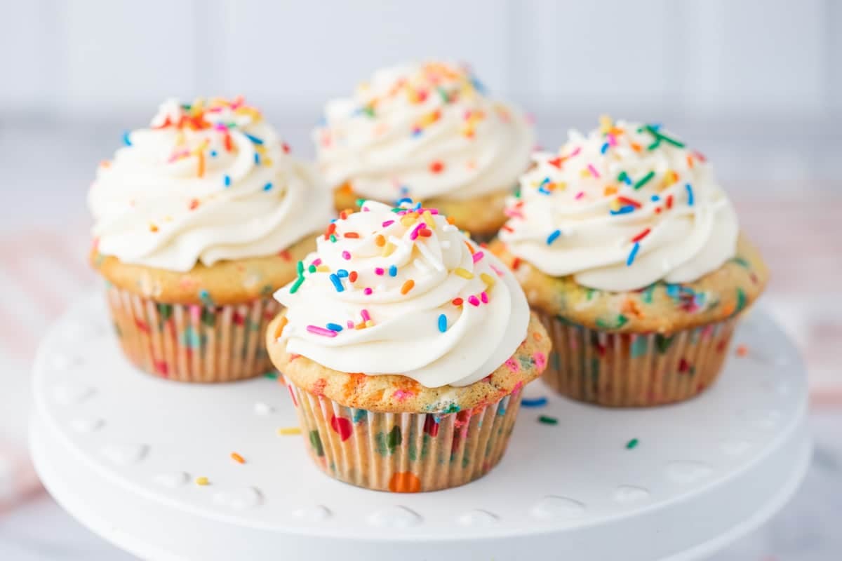Four cupcakes with white frosting and colorful sprinkles sit on a white cake stand.