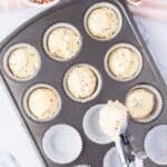 A muffin tin is filled with cupcake liners and scoops of sprinkle cupcake batter. A bowl of rainbow sprinkles and empty egg shells are nearby.