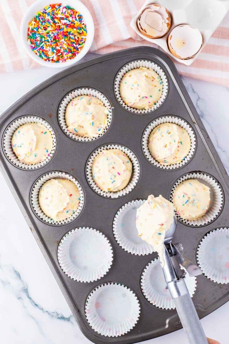A muffin tin is filled with cupcake liners and scoops of sprinkle cupcake batter. A bowl of rainbow sprinkles and empty egg shells are nearby.