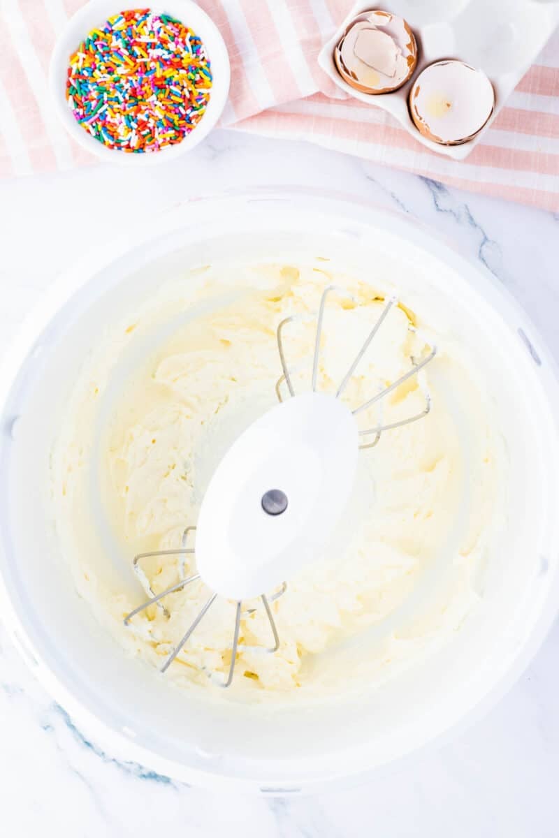 A mixing bowl with electric mixer beaters and creamy batter, next to a bowl of rainbow sprinkles and cracked eggshells on a striped cloth.