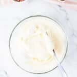 A glass bowl of white cake batter with a spatula, next to a bowl of rainbow sprinkles and empty eggshells on a striped cloth.