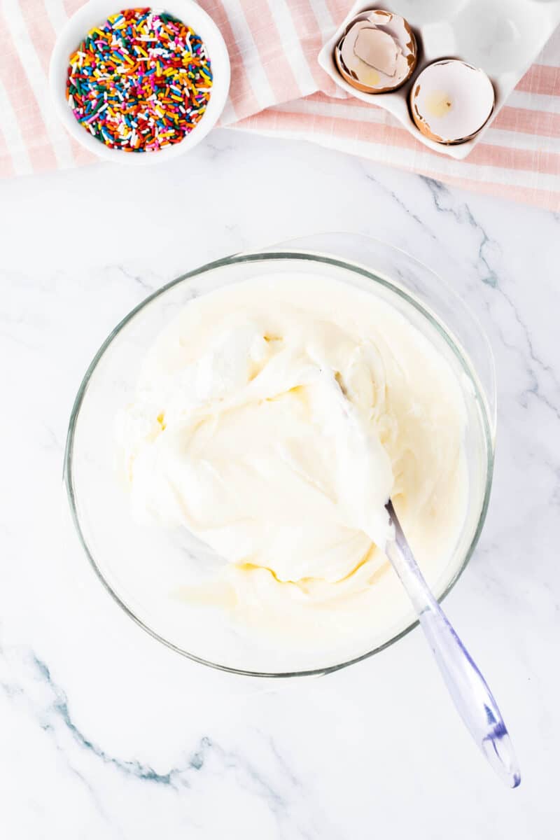 A glass bowl of white cake batter with a spatula, next to a bowl of rainbow sprinkles and empty eggshells on a striped cloth.