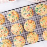 Twelve sprinkle cookies cooling on a wire rack, with a bowl of rainbow sprinkles and cracked eggshells on a striped cloth nearby.