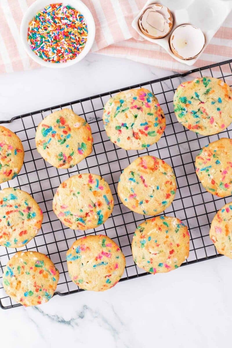 Twelve sprinkle cookies cooling on a wire rack, with a bowl of rainbow sprinkles and cracked eggshells on a striped cloth nearby.