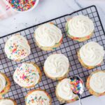 Twelve cupcakes with white frosting on a cooling rack, with some topped with colorful sprinkles; a spoon and a bowl of sprinkles are nearby.