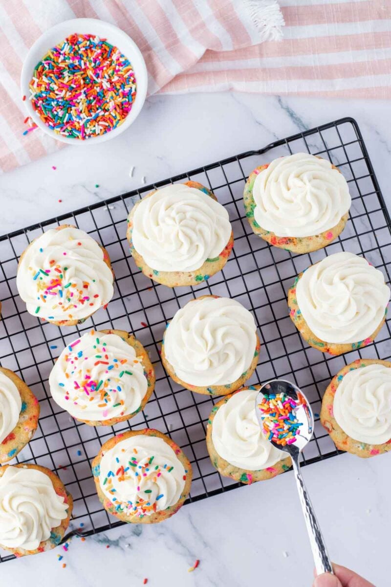 Twelve cupcakes with white frosting on a cooling rack, with some topped with colorful sprinkles; a spoon and a bowl of sprinkles are nearby.