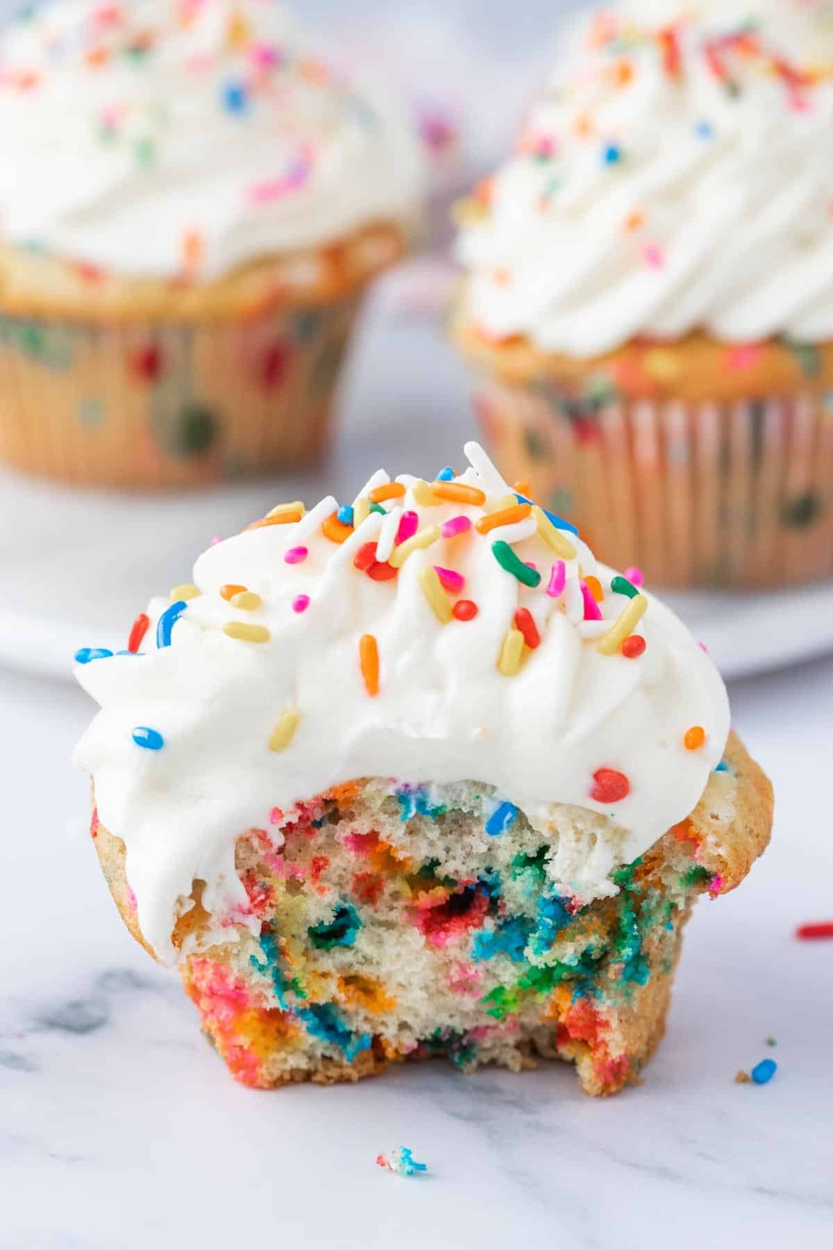 A bitten cupcake with white frosting and colorful sprinkles, showing a funfetti interior, with two whole cupcakes in the background.