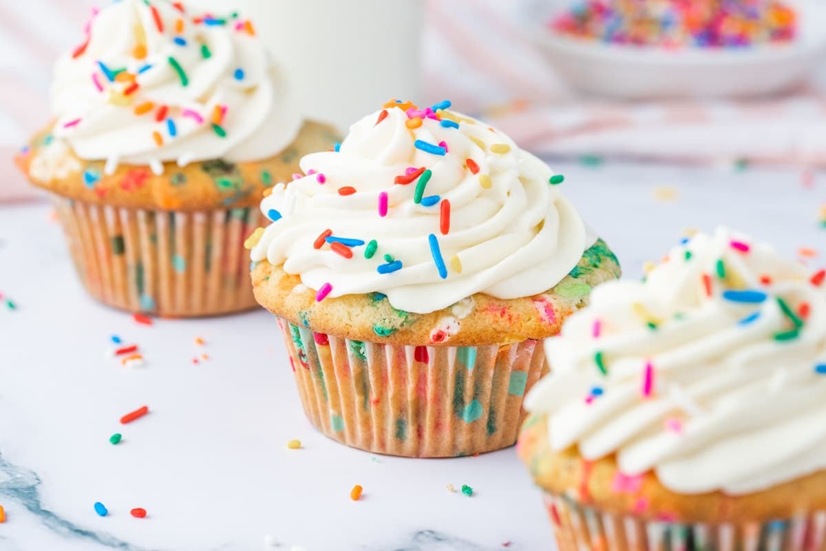 Three cupcakes with white frosting and colorful sprinkles are arranged on a white surface, with more sprinkles scattered around.