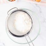 A sifter with flour over a glass bowl, two eggs in a carton, and a bowl of colorful sprinkles on a marble countertop.