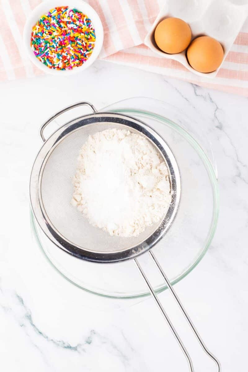 A sifter with flour over a glass bowl, two eggs in a carton, and a bowl of colorful sprinkles on a marble countertop.