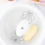 A mixing bowl with butter and sugar, two eggs in a carton, and a bowl of colorful sprinkles on a striped cloth beside it.