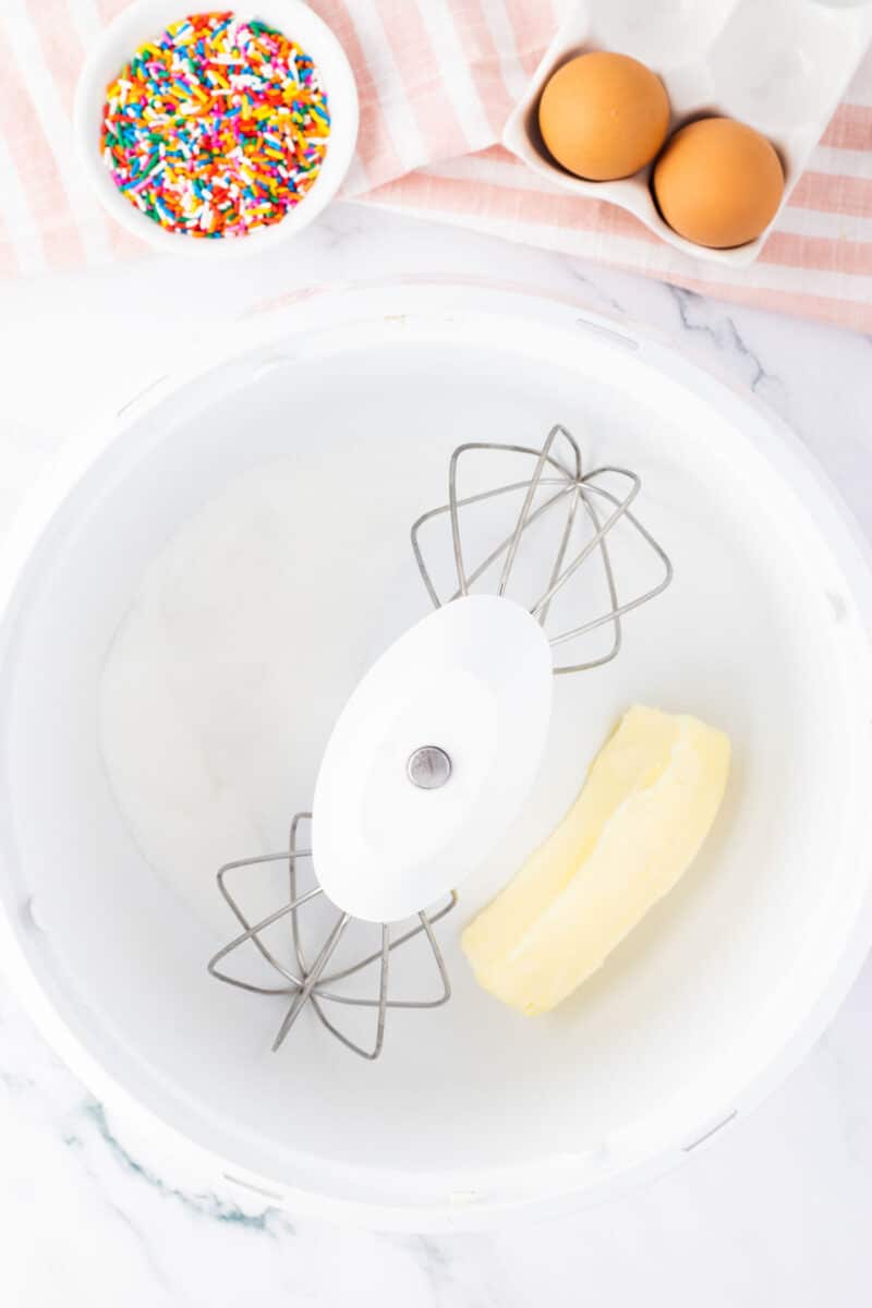 A mixing bowl with butter and sugar, two eggs in a carton, and a bowl of colorful sprinkles on a striped cloth beside it.