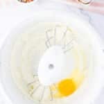 A mixing bowl with creamed mixture, an egg, and beaters. Nearby are a bowl of rainbow sprinkles, a cracked egg, and part of a pink striped towel.