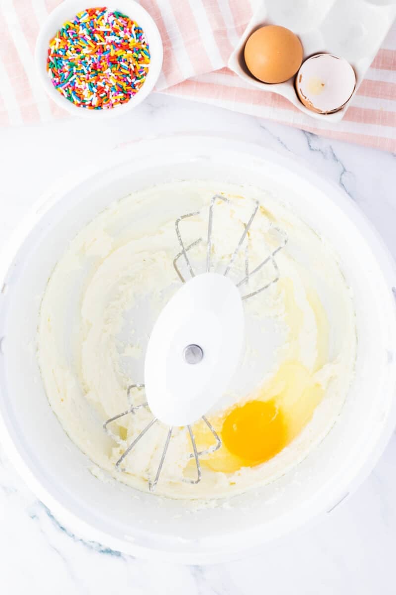 A mixing bowl with creamed mixture, an egg, and beaters. Nearby are a bowl of rainbow sprinkles, a cracked egg, and part of a pink striped towel.