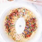 A mixing bowl with cake batter and colorful sprinkles, next to a bowl of sprinkles, cracked eggshells, and a striped towel on a marble surface.