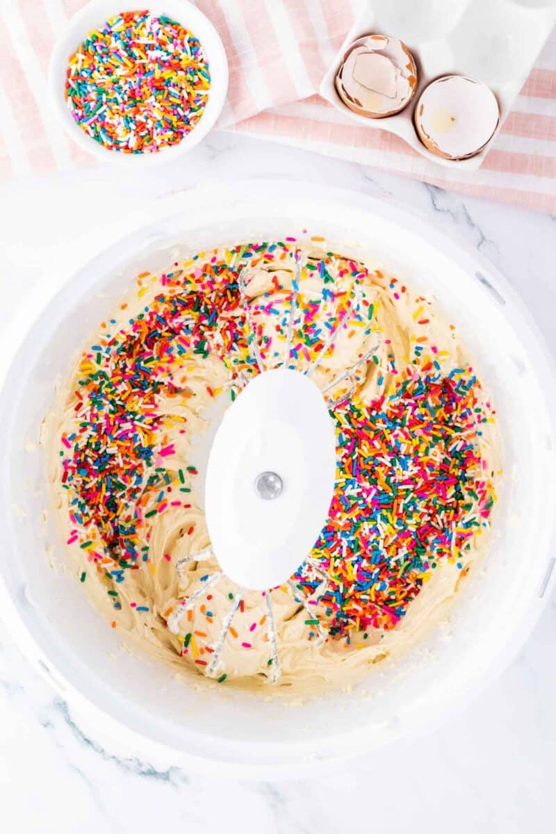 A mixing bowl with cake batter and colorful sprinkles, next to a bowl of sprinkles, cracked eggshells, and a striped towel on a marble surface.