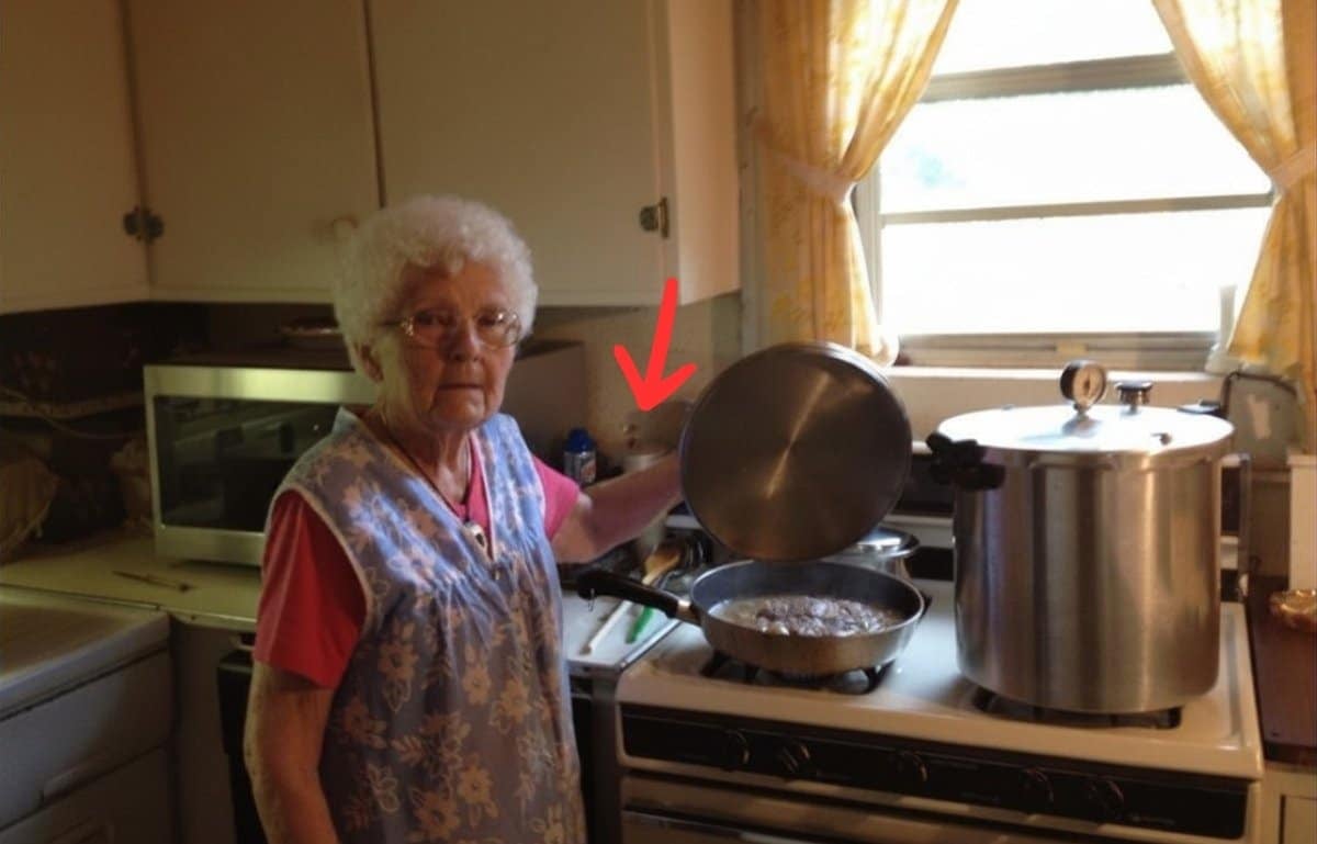 An elderly woman stands in a kitchen, holding a lid over a frying pan on the stove next to a pressure cooker, with an arrow pointing to the pan.