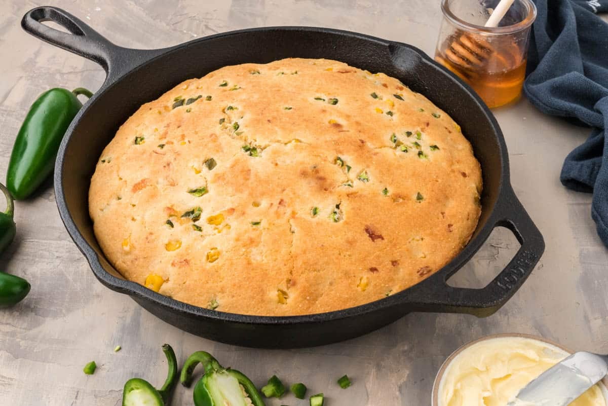 A skillet of baked cornbread topped with chopped jalapeños, shown with butter, sliced jalapeños, and a jar of honey nearby.
