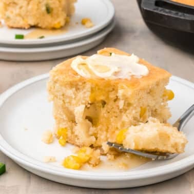 A square slice of cornbread with corn kernels, topped with butter, sits on a white plate with a forkful removed; a glass jar and jalapeño slices are nearby.