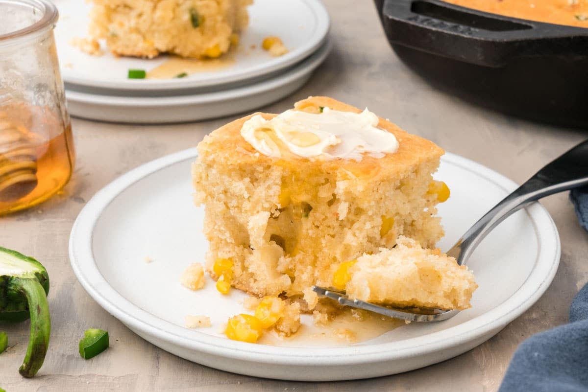 A square slice of cornbread with corn kernels, topped with butter, sits on a white plate with a forkful removed; a glass jar and jalapeño slices are nearby.