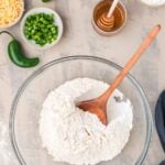 A glass bowl with flour mixture and a wooden spoon, surrounded by chopped jalapeños, shredded cheese, honey, sour cream, and whole jalapeños on a countertop.