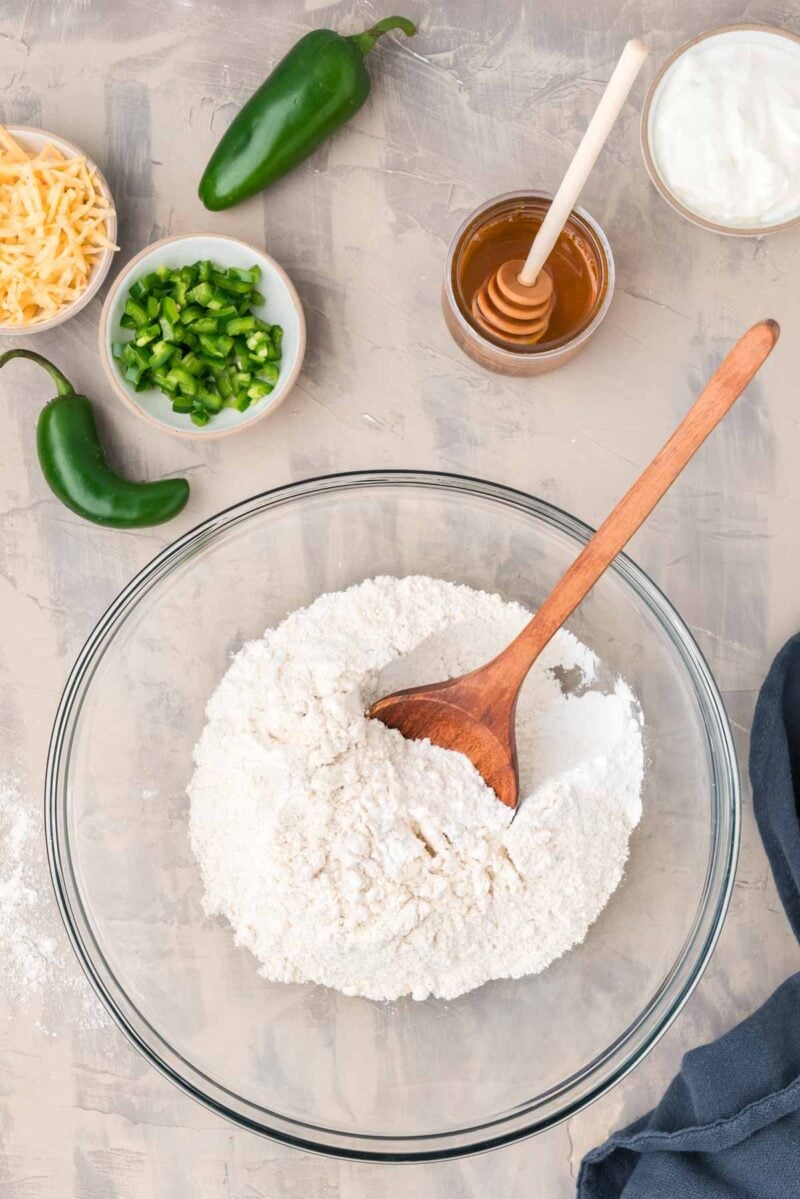 A glass bowl with flour mixture and a wooden spoon, surrounded by chopped jalapeños, shredded cheese, honey, sour cream, and whole jalapeños on a countertop.