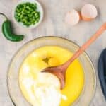 A glass bowl with eggs, melted butter, and milk being mixed with a wooden spoon, surrounded by corn, jalapeños, shredded cheese, and empty egg shells.