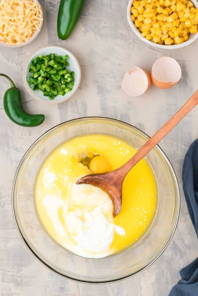 A glass bowl with eggs, melted butter, and milk being mixed with a wooden spoon, surrounded by corn, jalapeños, shredded cheese, and empty egg shells.