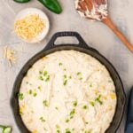 A cast iron skillet filled with cornbread batter topped with chopped jalapeños and corn, surrounded by jalapeños, shredded cheese, and a wooden spoon with batter.