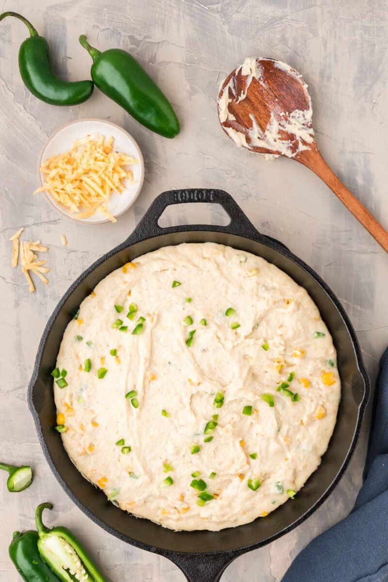 A cast iron skillet filled with cornbread batter topped with chopped jalapeños and corn, surrounded by jalapeños, shredded cheese, and a wooden spoon with batter.