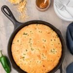 A round jalapeño cornbread in a cast iron skillet, surrounded by sliced jalapeños, shredded cheese, honey, plates, and cutlery on a gray surface.