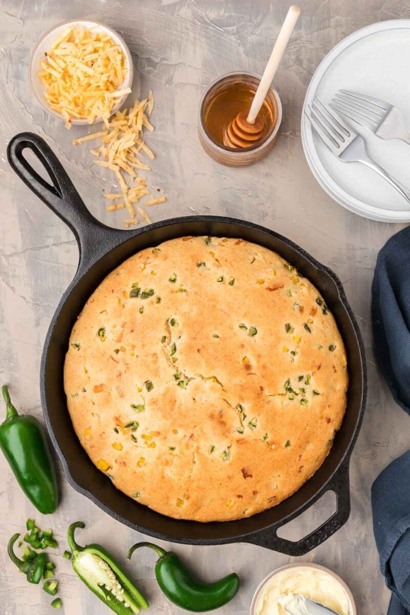 A round jalapeño cornbread in a cast iron skillet, surrounded by sliced jalapeños, shredded cheese, honey, plates, and cutlery on a gray surface.