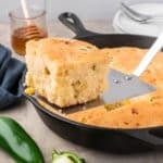 A slice of jalapeño cornbread is lifted from a cast iron skillet with a spatula; sliced jalapeños, honey, and plates are nearby.