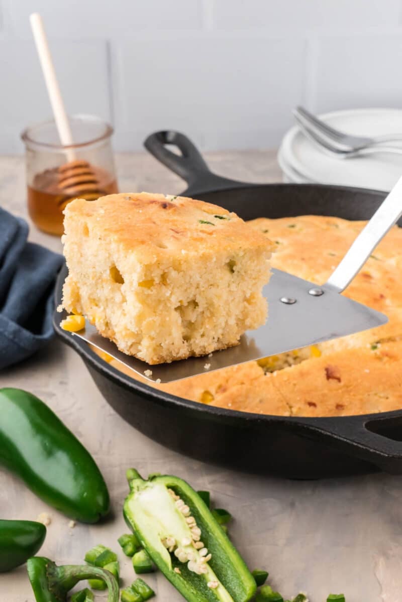 A slice of jalapeño cornbread is lifted from a cast iron skillet with a spatula; sliced jalapeños, honey, and plates are nearby.