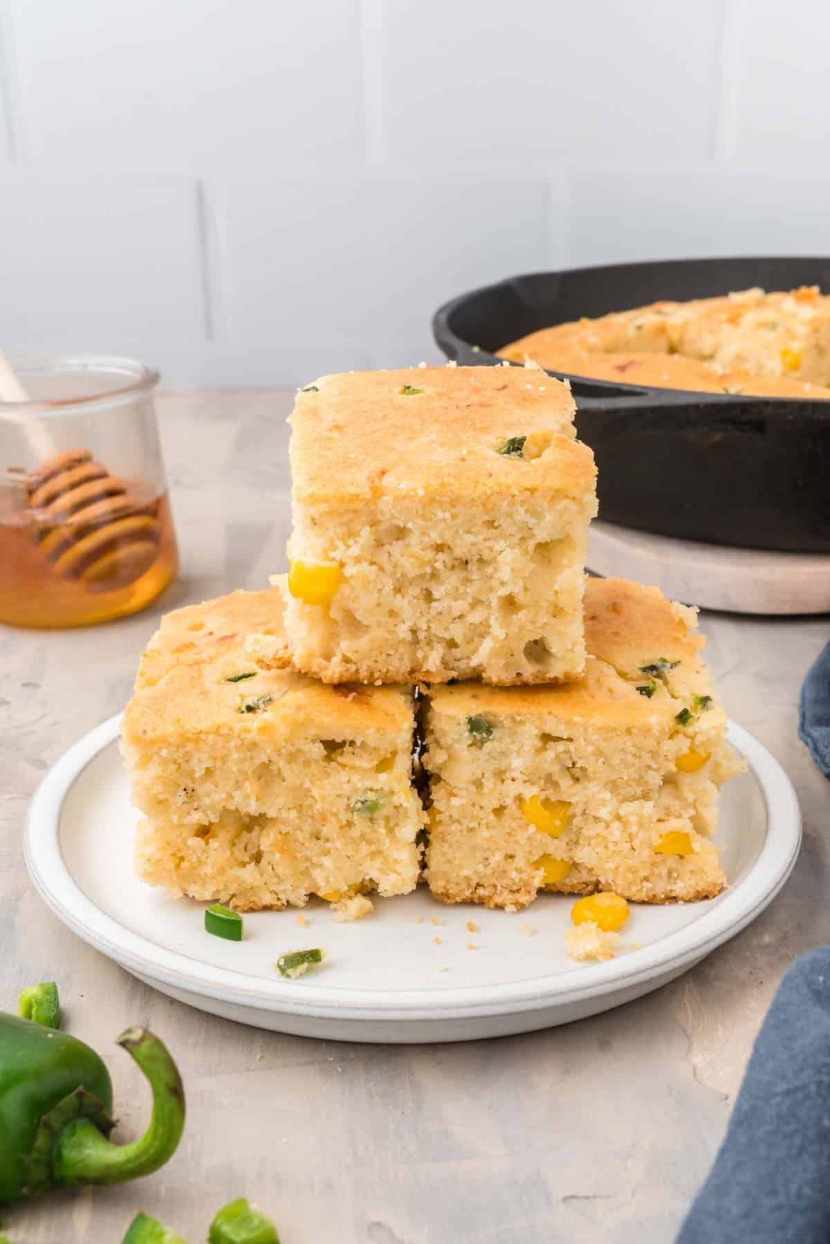 Three pieces of cornbread with corn and green pepper are stacked on a plate, with a skillet, honey jar, and jalapeño in the background.