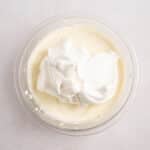 A glass bowl containing creamy white mixture with a dollop of whipped topping on a light-colored surface.