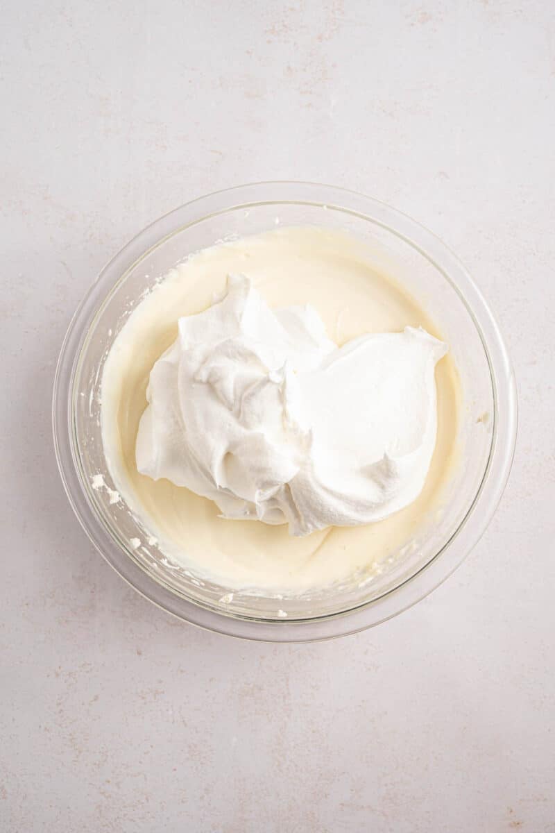 A glass bowl containing creamy white mixture with a dollop of whipped topping on a light-colored surface.
