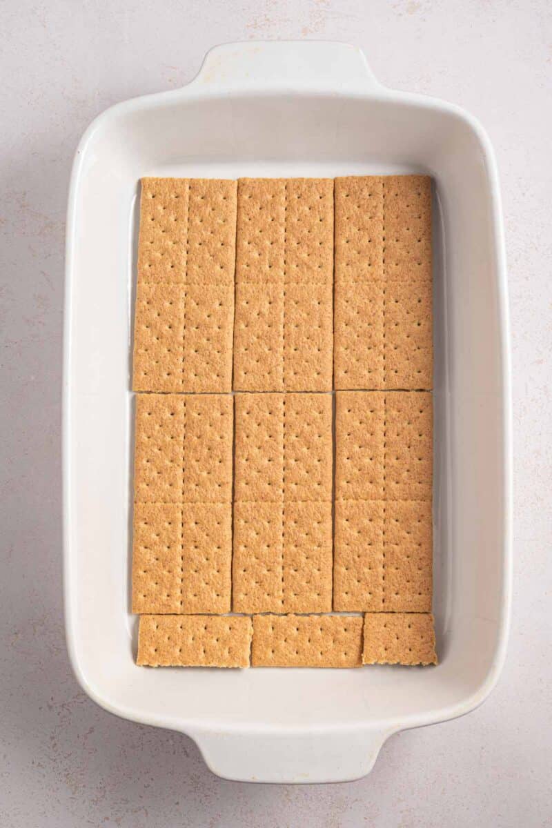 Rectangular graham crackers are neatly arranged in a single layer at the bottom of a white rectangular baking dish.