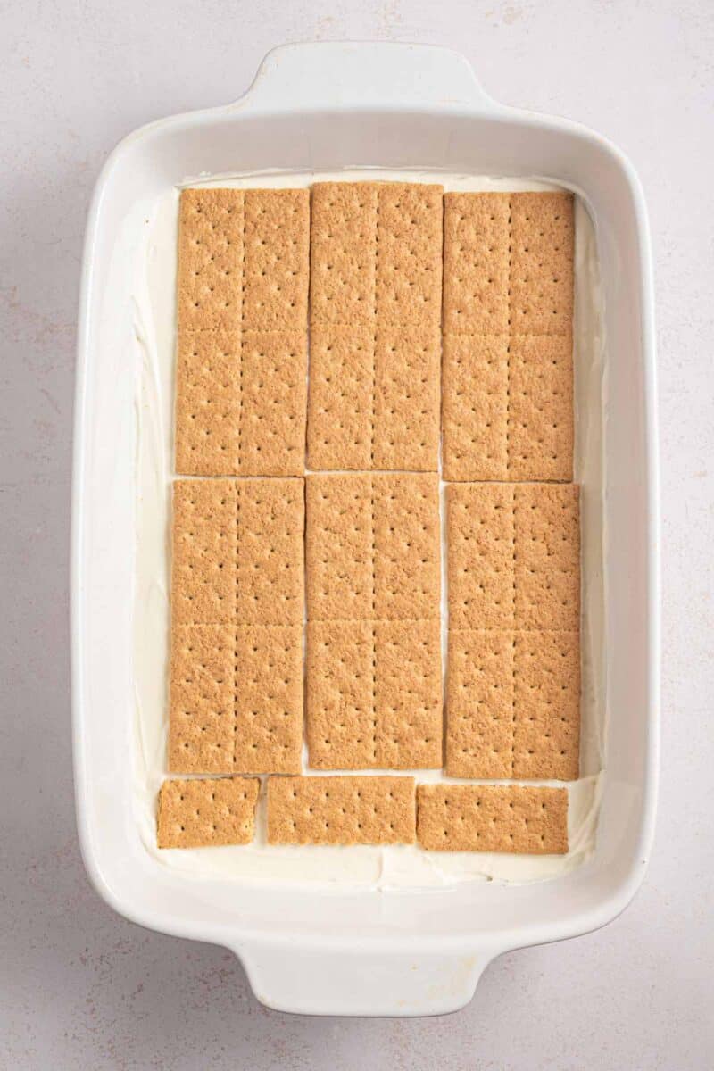 Rectangular baking dish with a layer of graham crackers on top of a white creamy base, placed on a light-colored surface.
