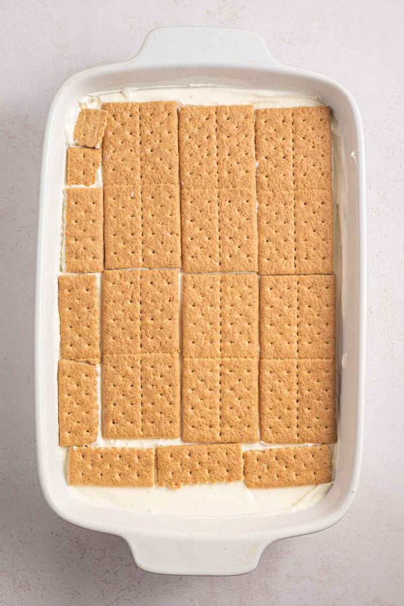 Rectangular baking dish with a layer of whole and broken graham crackers placed over a creamy white filling.