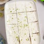 A white rectangular dish with sliced key lime icebox cake topped with whipped cream and lime zest, surrounded by lime halves and kitchen utensils.