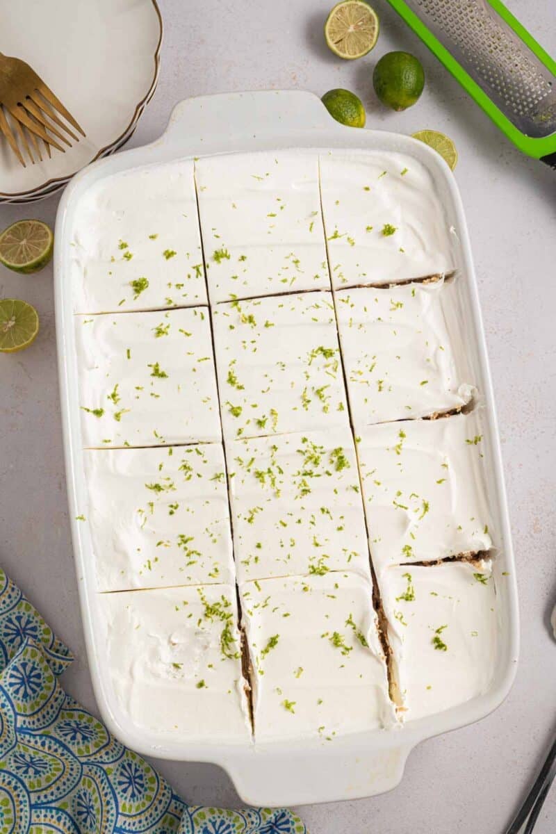 A white rectangular dish with sliced key lime icebox cake topped with whipped cream and lime zest, surrounded by lime halves and kitchen utensils.