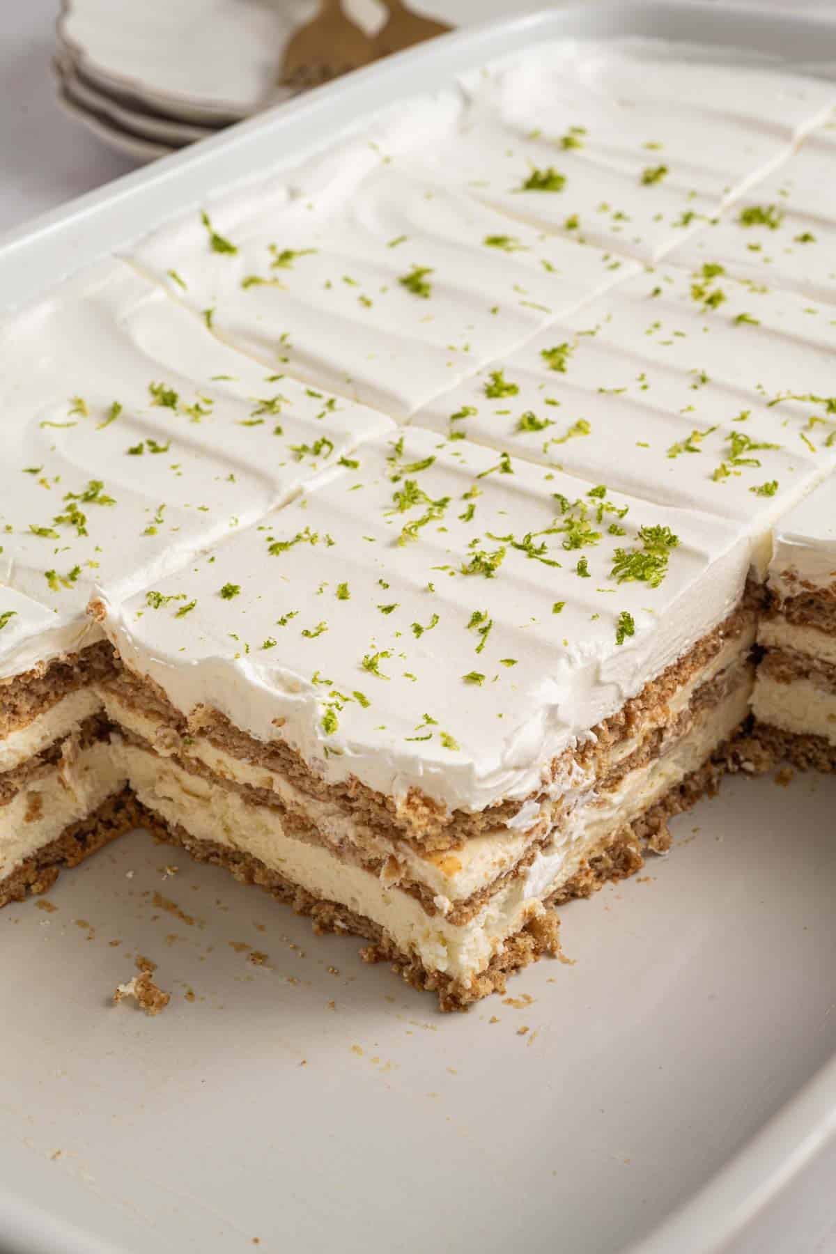 A rectangular dish of key lime icebox cake with whipped topping, graham crackers, and lime zest, with several pieces already served.