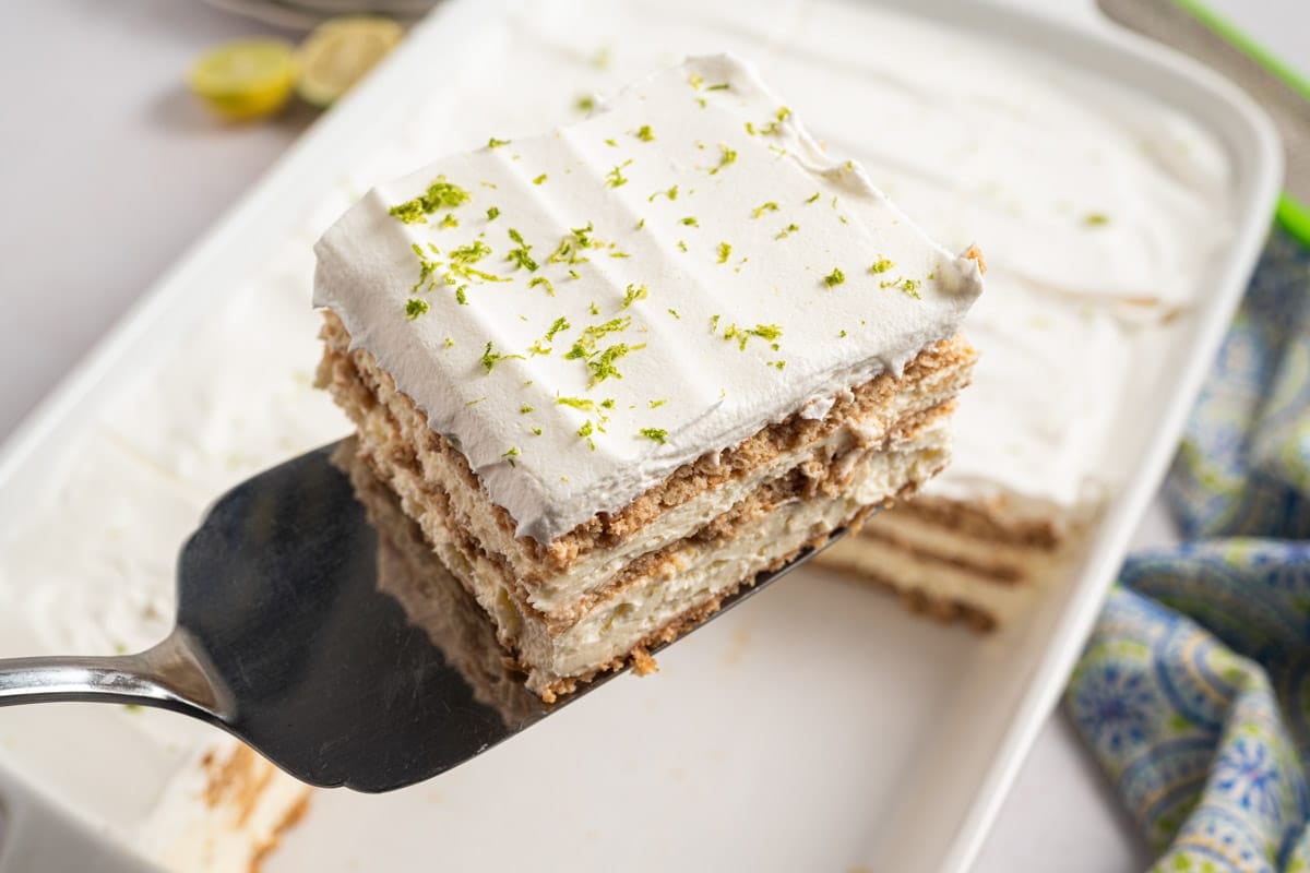 A spatula holds a square slice of key lime icebox cake with whipped topping and lime zest, above a white baking dish containing more of the dessert.