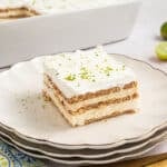 A slice of layered key lime icebox cake with cream and graham crackers is served on a plate, garnished with lime zest. Whole and halved limes, utensils, and a napkin are nearby.