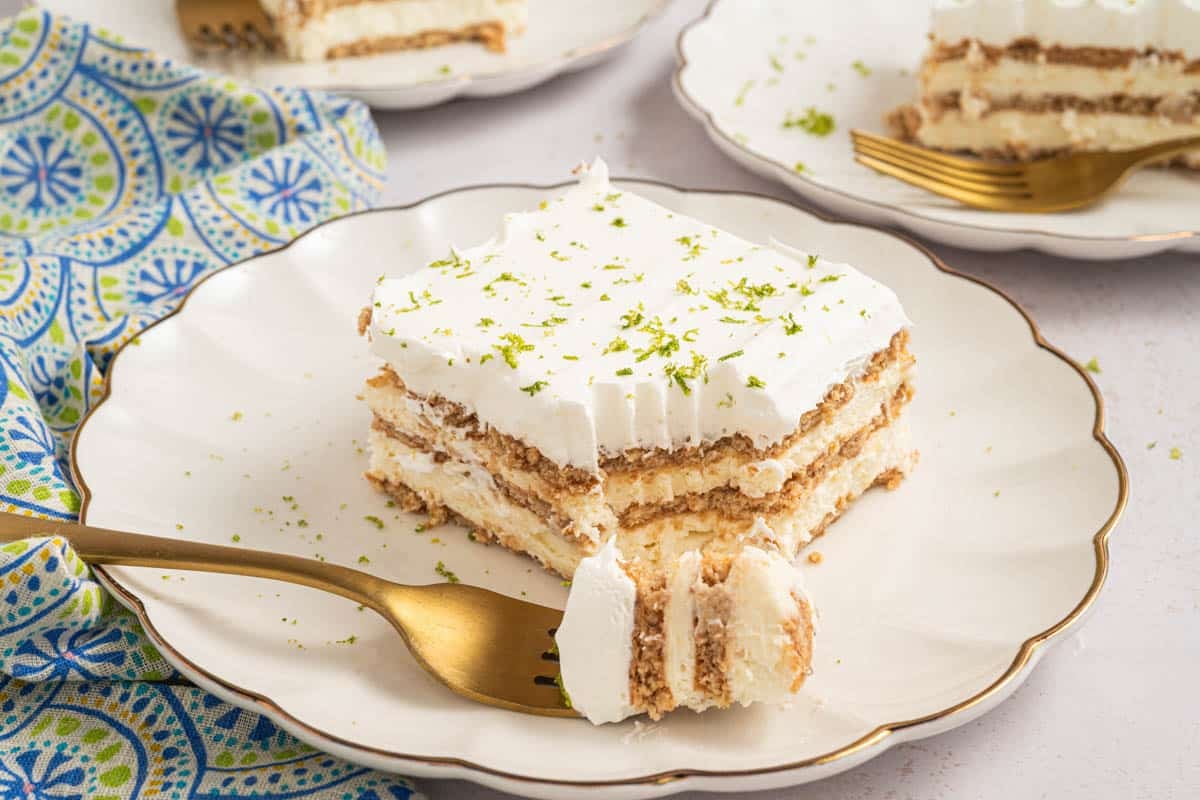 A slice of layered key lime icebox cake with cream and graham crackers on a white plate, topped with lime zest, with a gold fork holding a bite.